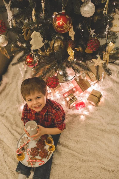 Boy laying under christmas tree with chocolate drink. — Stock Photo, Image