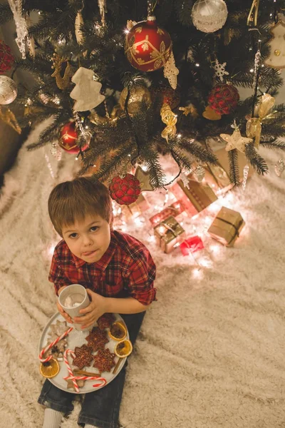 Boy laying under christmas tree with chocolate drink. — Stock Photo, Image