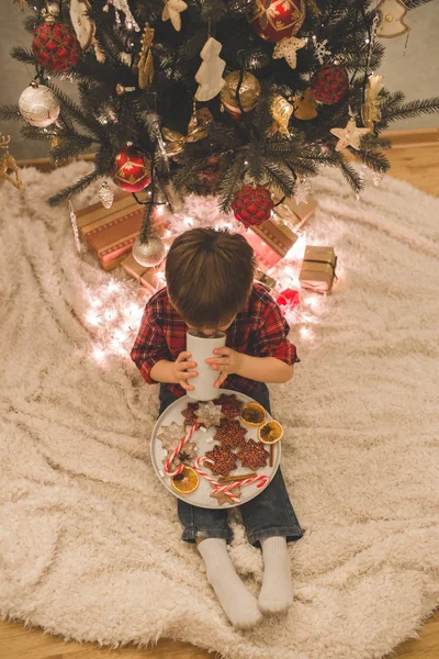 Boy laying under christmas tree with chocolate drink. — Stock Photo, Image