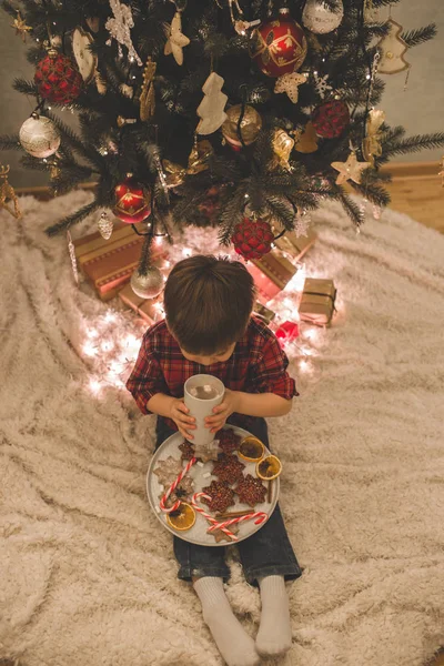 Boy laying under christmas tree with chocolate drink. — Stock Photo, Image