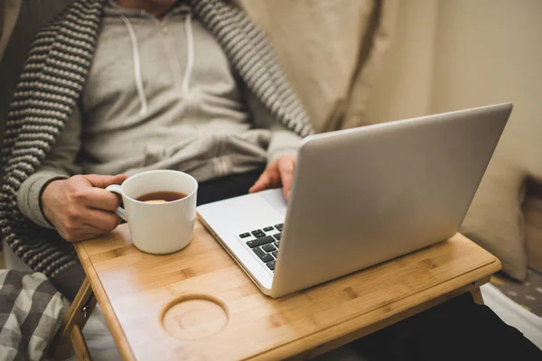 Jeune homme couché dans le mauvais avec ordinateur portable et tasse de thé . — Photo