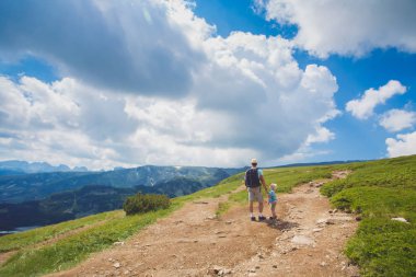 Baba ve oğul Rila mountains, Bulgaristan hiking. Bir gezgin ve küçük bir çocuk el ele tutuşarak yolda yürüyor. Arkadan Görünüm. büyük beyaz bulutlar üzerinde mavi bir gökyüzü