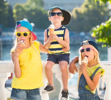 İki kız kardeş ve erkek kardeşleri vaiel bardağında dondurma yiyorlar. Ergenler şık giyinir. Sokak manzarasına karşı poz veren çocuklar