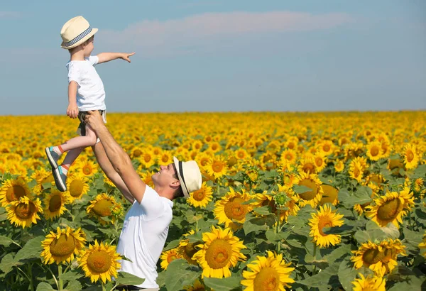 一个孩子和他的爸爸正在一个巨大的向日葵场玩耍 小男孩和男人穿着白色的T恤和帽子 爸爸把孩子抱在怀里 — 图库照片