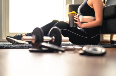 Spor ve ağırlık çalışmasından sonra protein ya da iyileşme içeceği içen kadın. Antrenman ve evde egzersiz. Spor giyim mağazasındaki kadın. Salaklar ve spor aletleri yere..