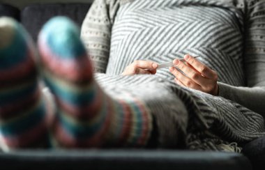 Grip ve soğuk algınlığı olan hasta bir kadın. Vücut ısısını termometreyle ölçüyor. Evdeki kanepede battaniyenin altında sıcak yün çoraplarla oturan hasta bir insan. Virüs, enfeksiyon, norovirüs veya hipotermi..