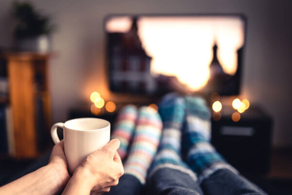 Couple drinking tea, hot chocolate, eggnog or mulled wine and watching tv in warm cozy woolen socks in winter. Woman holding cup of morning coffee in home living room. Sick people with flu or fever.
