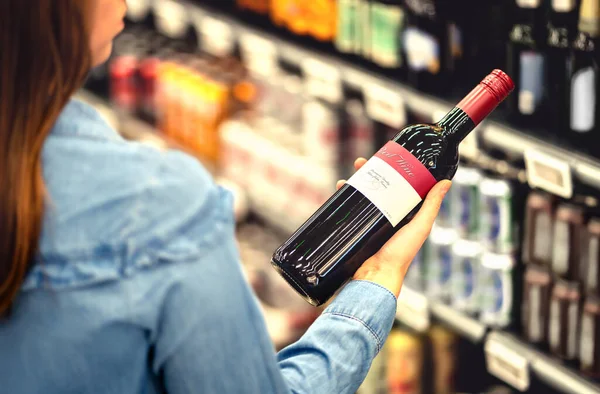 女人在超市的酒铺或酒类部阅读红葡萄酒瓶子的标签 架子上全是酒精饮料 持有和选择一瓶美乐或桑乔维斯葡萄酒的女顾客 — 图库照片