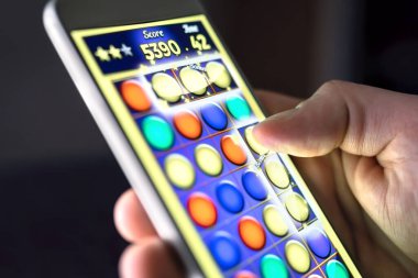 Akıllı telefon ekranında mobil oyun. Genç adam telefon uygulamasında bulmaca oynuyor. Oyuncu eğleniyor. Dijital oyun, eğlence ve video oyunu konsepti. Gece geç saatte tembel ya da bağımlı bir adam..