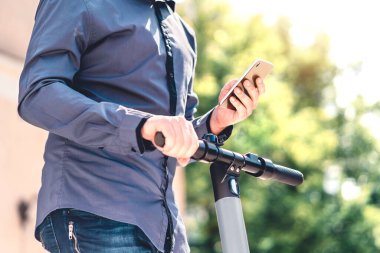 Elektrikli scooter ve cep telefonu. İş adamı, şehir caddesi ve parkında akıllı telefonlu bir araç kiralama servisi kullanıyor. Genç milenyum adamı yazın cep telefonu kiralayarak çevre taşımacılığı yapıyor.