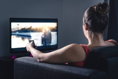 Kadın film izliyor ya da dizi seyrediyor. Filmi seçmek ya da kanalı değiştirmek için akıllı televizyon uzaktan kumandası kullanan kişi. Televizyon ekranında talep üzerine yayın veya video (VOD) servisi. Dijital eğlence.