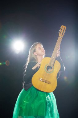 Yeşil elbiseli sevimli, uzun saçlı bir kız, parlak bir ışık altında, akustik gitarıyla solo bir konserde şarkı söylüyor.