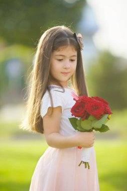 Bir buket gülü olan güzel küçük bir kız yeşil bir manzaranın arka planında duruyor. Yılın bu zamanı yaz..
