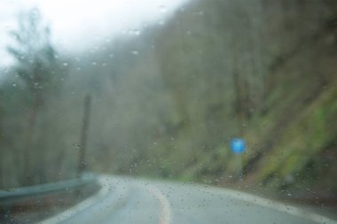 Yolun kenarındaki orman yollarının bulanık arkaplanı bulanıklaşmış. Arabanın ön camındaki yağmur damlalarına odaklan..