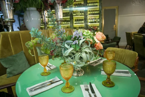 Стол на обед: столовые приборы, стаканы из желтого стекла и букет цветов в металлической вазе на ноге — стоковое фото