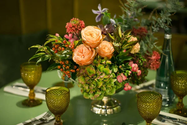 Table for lunch: cutlery, glasses of yellow glass and a bouquet of flowers in a metal vase on the leg — Stock Photo, Image