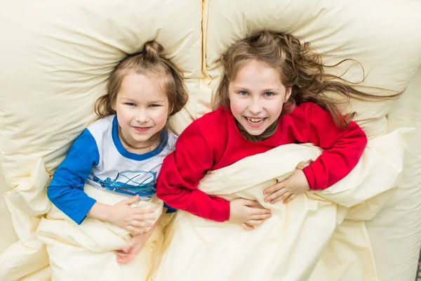 Lachen in rode pyjama meisje en een jongen in blauwe pyjama's liggen op het bed — Stockfoto