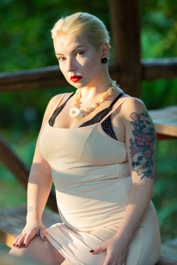 Bej elbiseli sarışın ormanda bir bankta oturuyor.