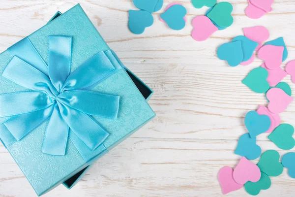 Blue gift box and a pile of paper hearts of pastel colors on the white wooden background (flat lay, copy space in the center for your text)