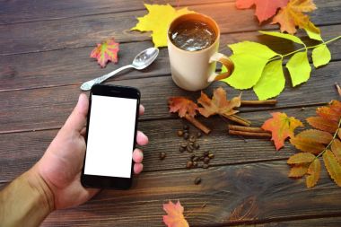 Bir adam bir telefon sol elinde tutuyor ve bir fincan kahve ve sonbahar bir natürmort arka plan beyaz ekran mesaj okuma ahşap bir masaya bırakır