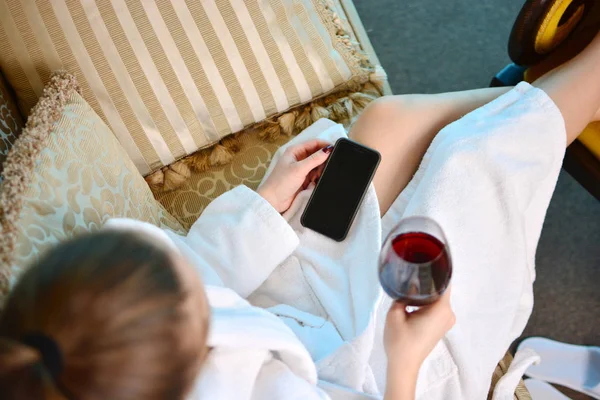 スマート フォンとワインで白いバスローブの女性はホテルのベッドでリラックス — ストック写真