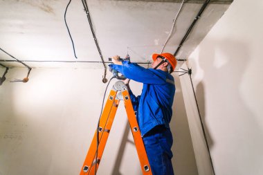 Sert şapkalı ve üniformalı elektrikçi, merdivende duruyor ve çekiç matkabıyla çalışıyor.