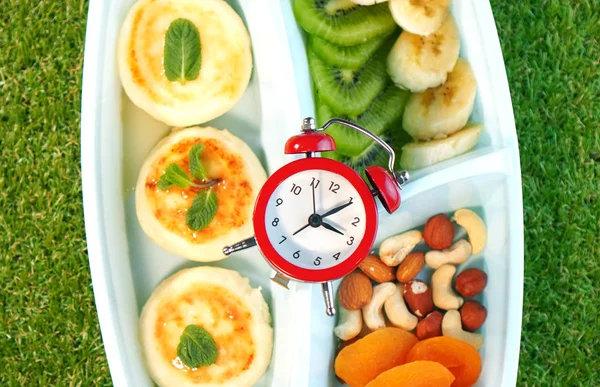 It\'s time to lunch break. Lunch box with cheesecakes, honey, kiwi, bananas, nuts and dried apricots. Clock lie on top.