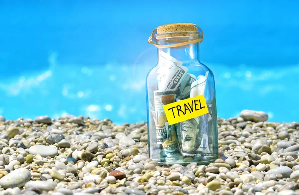 Summer vacation. Time to collect money for travel. Concept with glass money box full of dollars signed Travel.  Travel money box standing on a beach. Sea waves on a backdrop.