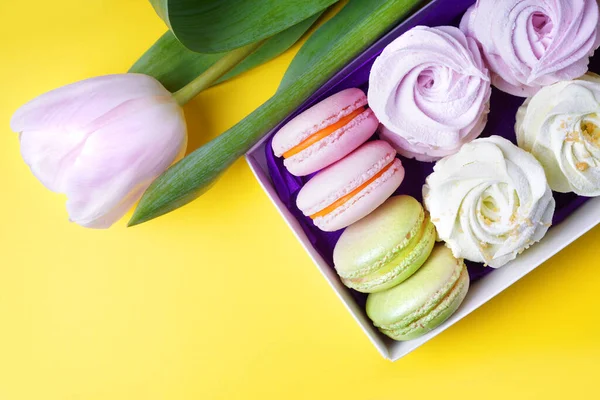 Box with macaroon and marshmallow cakes on a yellow background. Tulip lying near the box with sweets. Copy space. Top view.