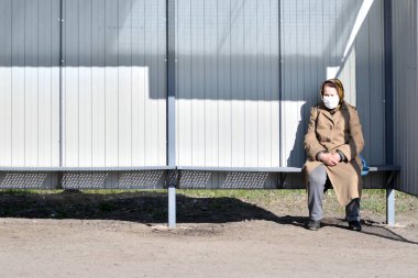 Kharkiv, Ukrayna - 5 Nisan 2020: Coronavirus ve risk altındaki insanlar. Tıbbi maskeli yaşlı bir kadın otobüs durağında tek başına oturuyor.