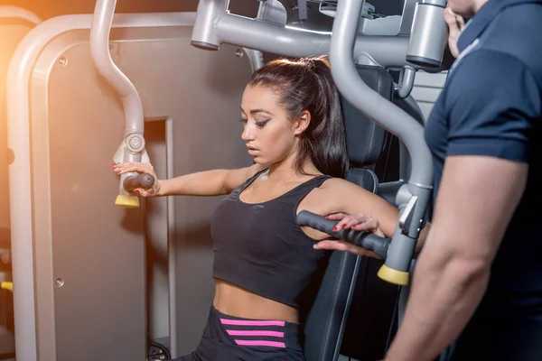 Sport, fitness, travail d'équipe et concept de personnes - jeune femme flexion des muscles sur la machine de gymnastique et entraîneur personnel avec presse-papiers — Photo