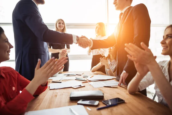 Benvenuto a bordo! Due uomini belli stringono la mano con il sorriso mentre si siedono sul divano in ufficio con i loro colleghi — Foto Stock