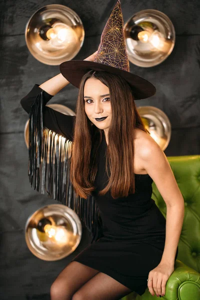 Mujer joven gótica feliz en traje de Halloween bruja con sombrero sentado en la silla — Foto de Stock