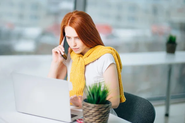 Mujer de negocios sentada en la oficina en el escritorio, mirando portátil y utiliza el teléfono inteligente —  Fotos de Stock