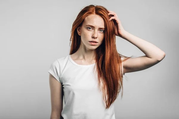 Jovem menina ruiva bonita com sardas olhando para a câmera sorrindo tocando o cabelo — Fotografia de Stock