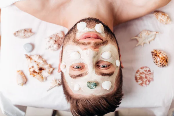 Relajante hombre en el salón de spa con máscara de conchas que ponen en la toalla blanca — Foto de Stock