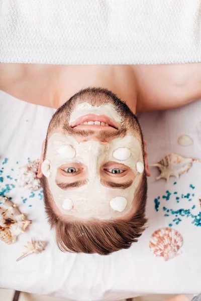 Relajante hombre en el salón de spa con máscara de conchas que ponen en la toalla blanca — Foto de Stock