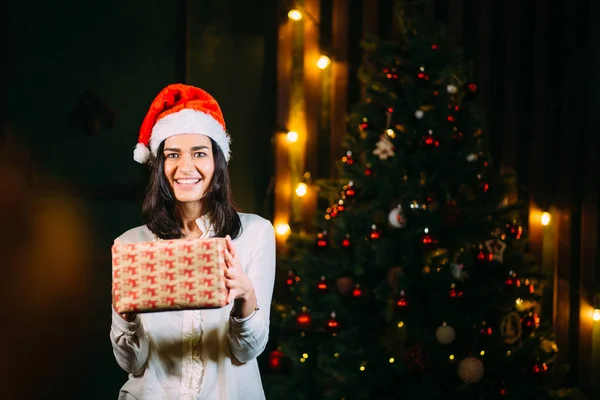 女孩在圣诞老人帽子附近在圣诞树与礼物 — 图库照片
