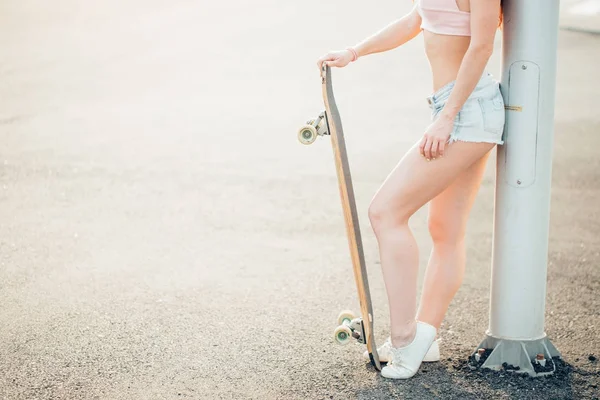 Menina na moda urbana com longboard posando ao ar livre na estrada ao pôr do sol . — Fotografia de Stock