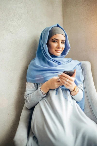 Retrato de mujer musulmana feliz usando el teléfono móvil mientras está sentado en un sofá — Foto de Stock