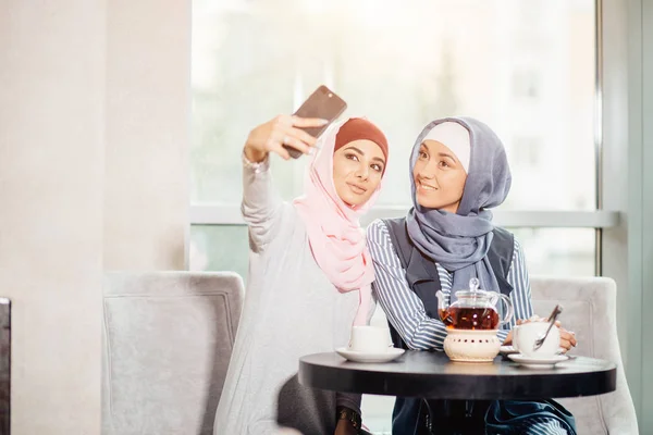 Two happy young muslim woman take self portrait with handphone at cafe — Stock Photo, Image