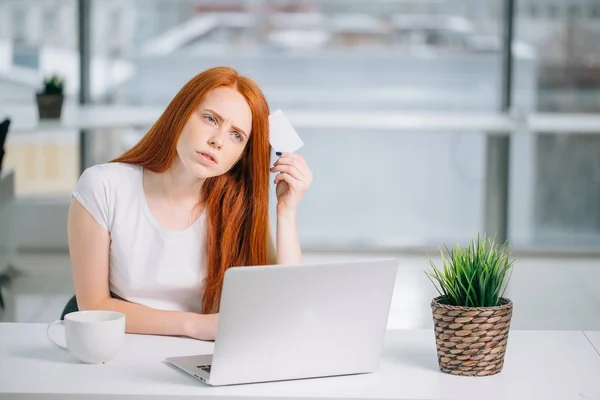 Pensando mulher de negócios sentada à mesa com laptop, segurando cartão de crédito vazio — Fotografia de Stock