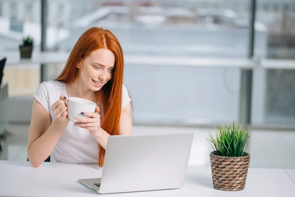 Счастливая улыбчивая рыжая женщина, работающая с ноутбуком и пьющая кофе — стоковое фото