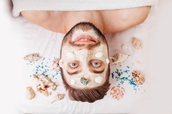 Relaxing man in spa salon with mask from shells laying on white towel — Stock Photo, Image