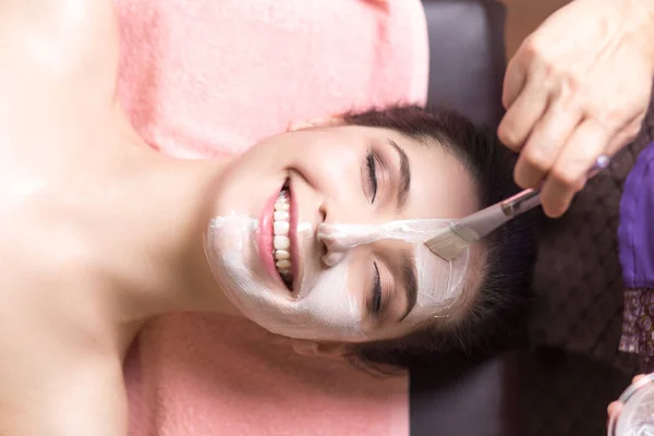 woman lying and cosmetologist applying facial mask by brush in spa