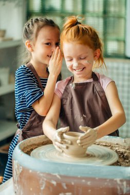 two girls sculpts from clay. one child whispers from her friend clipart