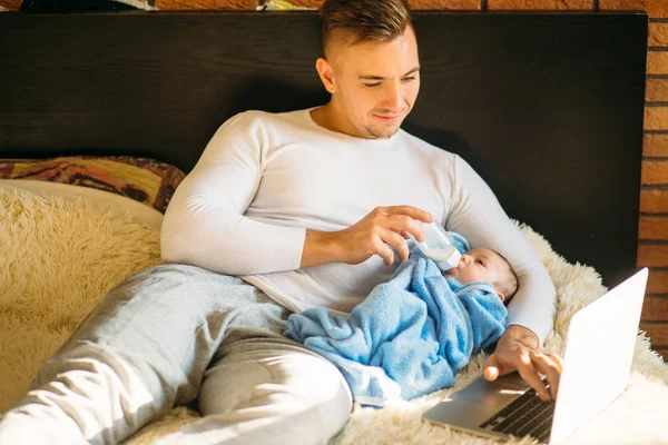 Hombre alimentando pequeño bebé mientras está acostado en la cama y trabajando en el ordenador portátil —  Fotos de Stock