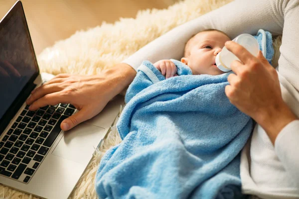 Hombre alimentando pequeño bebé mientras está acostado en la cama y trabajando en el ordenador portátil —  Fotos de Stock