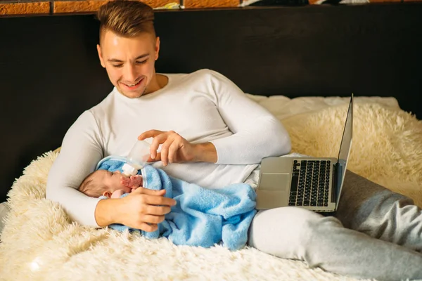 Muže, který krmí děťátko ležící na posteli a práci na notebooku — Stock fotografie