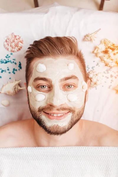 Relajante hombre en el salón de spa con máscara de conchas que ponen en la toalla blanca — Foto de Stock